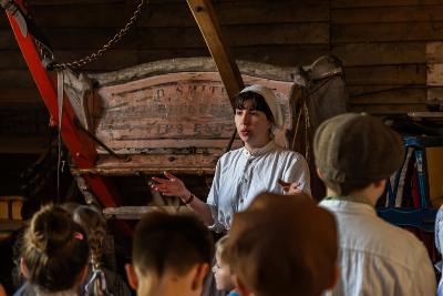 Photograph of museum worker, in historical costume, standing next to old machinery and speaking to children